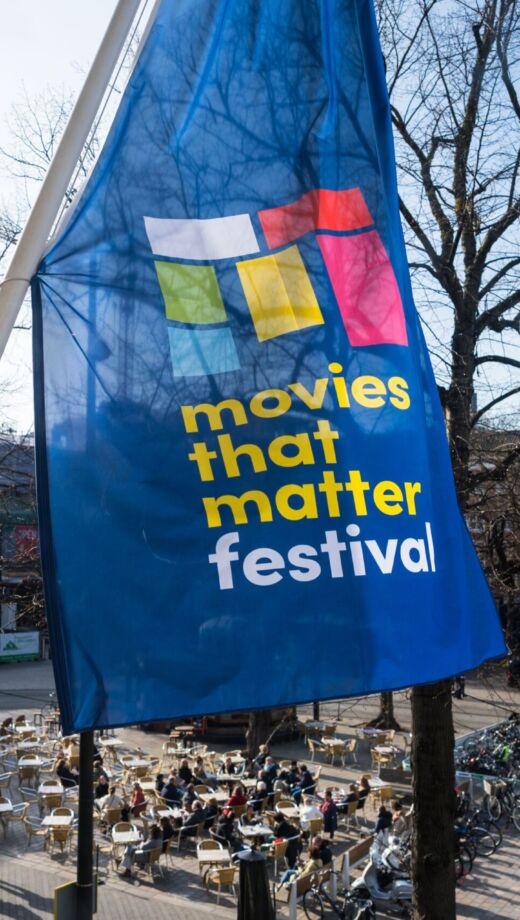 Blauwe vlag met gekleurd Movies that Matter M logo en geel/witte tekst Movies that Matter Festival. Op achtergrond is Buitenhof in Den Haag te zien.