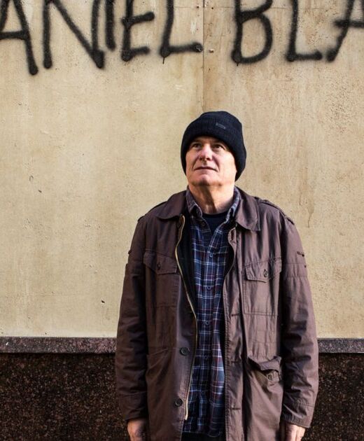 Foto van I, Daniel Blake. Oudere witte man, met een zwarte muts op en jas aan, staat voor een grijze muur met graffiti.