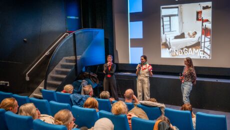 Drie sprekers staan voor een publiek in een bioscoopzaal met blauwe stoelen. Op de achtergrond is een groot scherm te zien met de titel van een film genaamd The Mind Game en informatie over het festival, dat plaatsvindt van 22 tot 30 maart 2024. De zaal zit vol met aandachtig luisterende bezoekers.