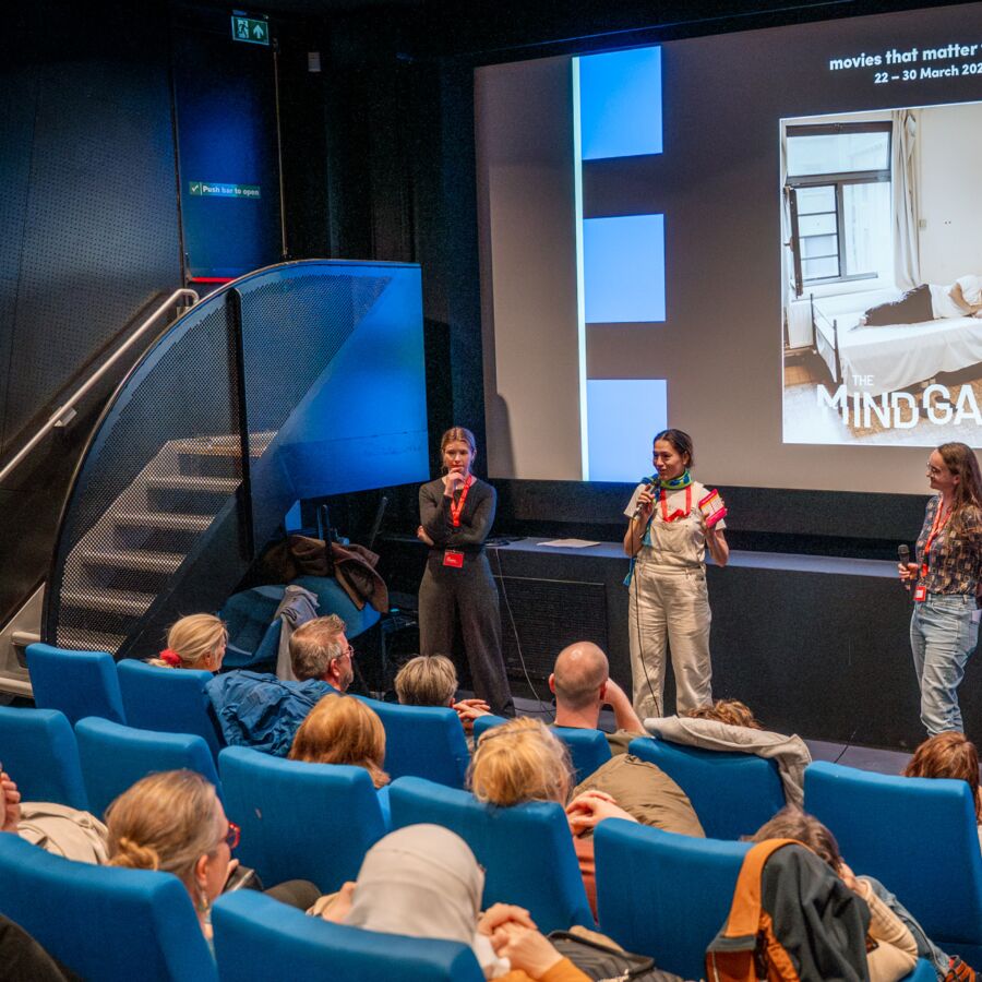 Drie sprekers staan voor een publiek in een bioscoopzaal met blauwe stoelen. Op de achtergrond is een groot scherm te zien met de titel van een film genaamd The Mind Game en informatie over het festival, dat plaatsvindt van 22 tot 30 maart 2024. De zaal zit vol met aandachtig luisterende bezoekers.