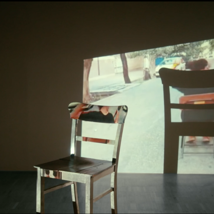 A white chair in front of a white wall, on which is projected an image of a woman turned away on a red chair.