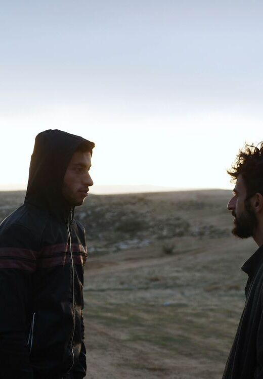 Een afbeelding uit No Other Land waarin twee mannen elkaar aankijken tegen de achtergrond van een landschap.