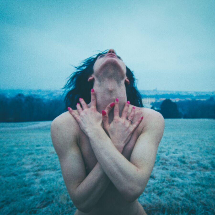 Person naked in an open field crosses arms across chest to throat with head tilted backwards