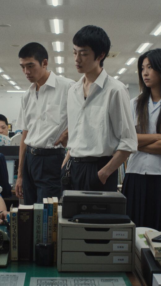 Group of students standing around a seated school staff member inside an office room.