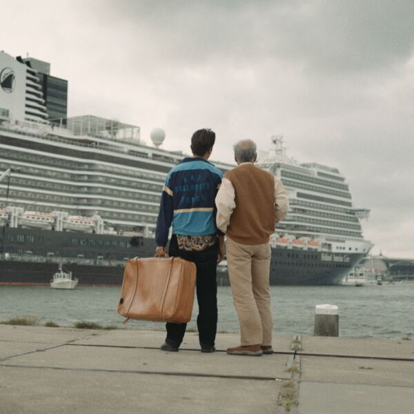 Twee mannen, rechts jong met een bruine koffer in zijn hand, links oud, kijkend over een kade, naar een groot cruiseschip.