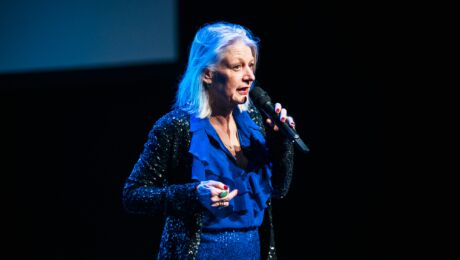 Woman with grey hair on a stage with a blue glitter suit and a microphone in hand.