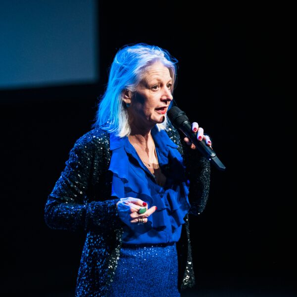 Vrouw met grijs haar en blauw glitter pak, staat op een podium sprekend met een microfoon in haar hand.