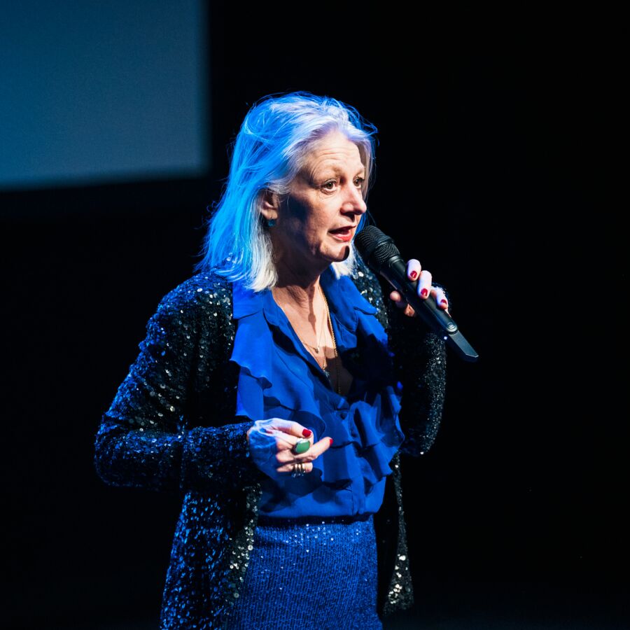 Woman with grey hair on a stage with a blue glitter suit and a microphone in hand.