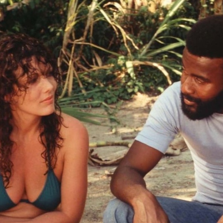 Een witte vrouw met donker krullend haar in bikini zit in de groene buitenlucht naast een donkere man met afro en baard, in grijs t-shirt en spijkerbroek.