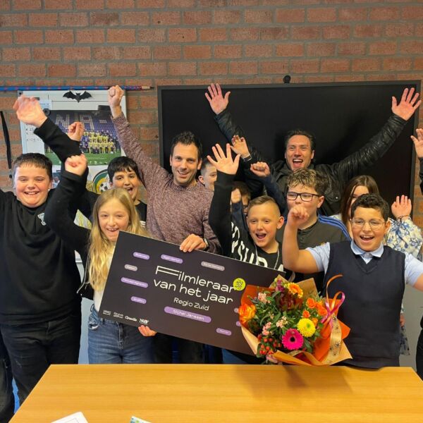 De winnaar van de Filmleraar van het Jaar verkiezing staat tussen zijn feestvierende leerlingen met een boeket en cheque.