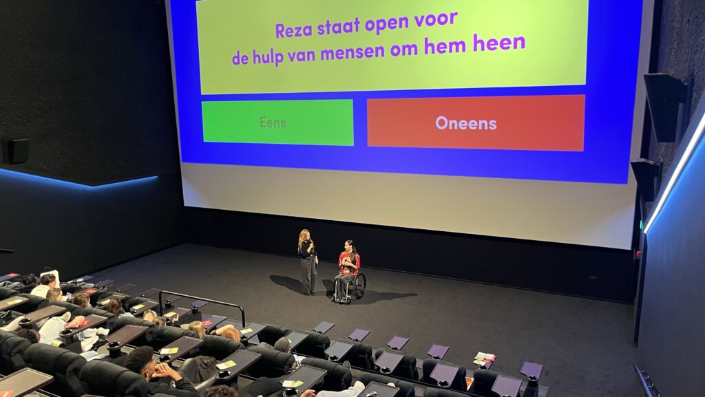 Leerlingen bekijken in de bioscoop een film en luisteren naar een spreker en een rolstoelgebruiker tijdens een nagesprek. Op het scherm staat een stelling over Reza en de hulp van anderen.